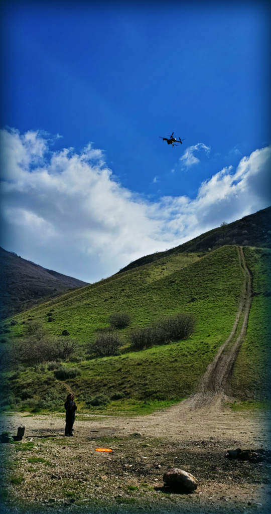 Drone flying in a cloudy sky above green hills. Pilot watching from below.